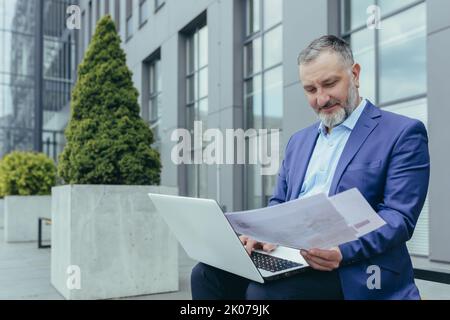 Senior stattlich erfolgreicher männlicher Ingenieur Architekt in Suit arbeitet mit Laptop, Dokumenten, Plan, Zeichnungen. Sitzt auf einer Bank in der Nähe des Bürozentrums, hält Papiere, schaut sich um, lächelt. Stockfoto
