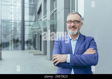 Porträt von älteren grauhaarigen schönen erfolgreichen erfolgreichen Geschäftsmann Mann in Brille und Anzug. Er steht auf der Straße in der Nähe des Bürozentrums, faltete seine Arme vor sich, blickt auf die Kamera und lächelt. Stockfoto