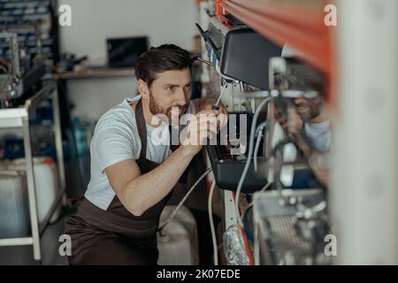 Lächelnder junger Mann, der in einer Werkstatt die Kaffeemaschine mit einem Schraubendreher repariert Stockfoto