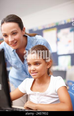 Sie verfügen über gute Computerkenntnisse. Eine attraktive junge Lehrerin, die einem niedlichen Schulmädchen im Computerunterricht hilft. Stockfoto