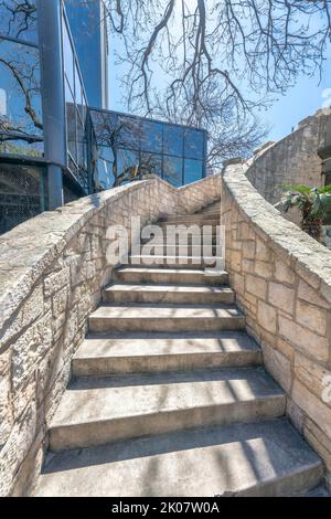Außentreppe mit sonnendurchfluteten Betonstufen und Steingeländern an einem sonnigen Tag. Blick vom unteren Ende einer Parktreppe mit Glasgebäude und blauen s Stockfoto
