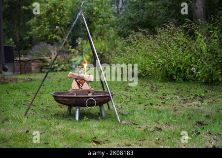 Gestapeltes Brennholz in einer Feuerschale brennt mit Flammen und raucht unter einem Dreibeingrill in einem Garten, Vorbereitung für eine Grillparty, Kopierplatz, ausgewählt Stockfoto