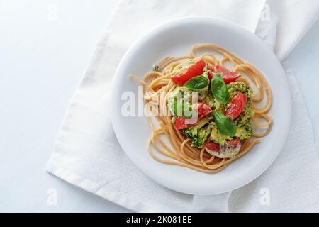 Vollkorn-Spaghetti mit Brokkoli-Sahnesauce, Tomate und Basilikum auf weißem Teller garnieren, gesunde vegetarische Mahlzeit, Kopierraum, Draufsicht von oben, Stockfoto