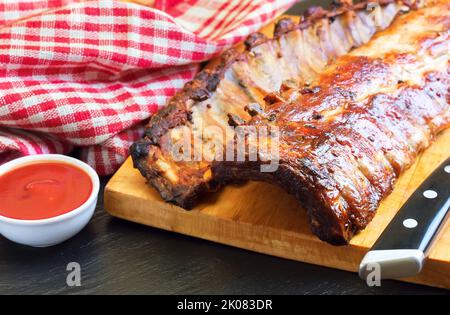 Gegrillte Schweineribs auf einem Holzschneidebrett mit Küchenmesser und Tomatenketchup Stockfoto