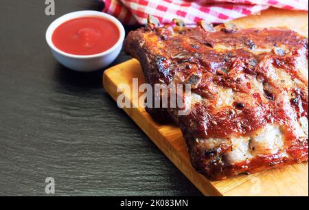 Gegrillte Schweinefleischrippen auf einem Holzschneidebrett mit Tomatenketchup auf schwarzer Schieferfläche Stockfoto