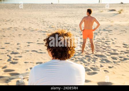 Zurück gedreht Blick weg zwei junge Männer auf sandigen Sommerstrand. Lockiges Haar Mann sitzt, ein anderer Mann ist Rettungsschwimmer in roten Badeshorts. Stockfoto