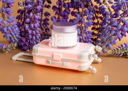 Feuchtigkeitsspendende Creme im Glasglas mit Schraubverschluss aus Metall, kleinem rosa Reisekoffer-Spielzeug und lila Lupine-Blüten viel auf braunem Hintergrund. Mockup, Fron Stockfoto
