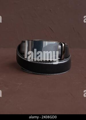 Aufgerollter schwarzer modischer Herren Ledergürtel mit automatischer Schnalle auf braunem Hintergrund. Männliches Zubehör. Vorderansicht, vertikales Foto. Stockfoto