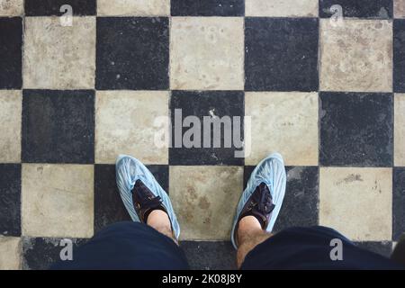 Mann mit blauen Schuhüberzügen, die über Stiefeln auf Fliesen getragen werden. Beine in Einweg-Schuhüberzügen vor dem Hintergrund des Bodens mit Fliesen bedeckt. Stockfoto