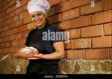 Schöne weibliche Bäckerin posiert mit einem traditionellen hausgemachten Sauerteig Weizenbrot vor rotem Backstein Wand Hintergrund Stockfoto