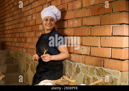 Porträt einer lächelnden hübschen Frau, einer Bäckerkonditorin, die mit einem Weizenspachtelchen vor rotem Backsteinmauer-Hintergrund posiert Stockfoto