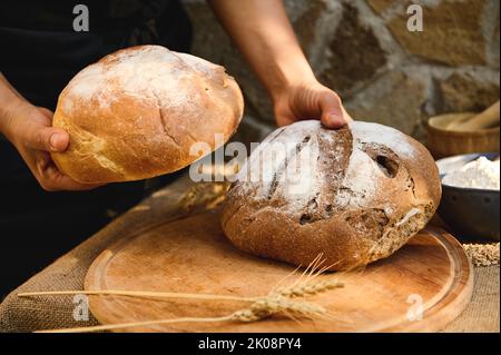 Details: baker Hände halten Laibe Vollkornsauerteig Brot, über einem hölzernen Schneidebrett mit Weizenspikelets Stockfoto