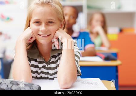 Dafür zu sorgen, dass ihre Zukunft so hell wie sie sein wird. Süßes junges Mädchen, das in der Klasse an ihrem Schreibtisch sitzt und einen anlächelt - Copyspace. Stockfoto