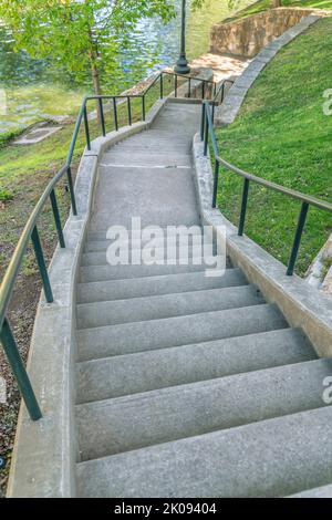 Eine Freitreppe in einem Park führt durch einen malerischen Park mit plätscherndem Wasser. Blick von der Spitze einer Beton-Außentreppe mit Metallgeländern inmitten sc Stockfoto