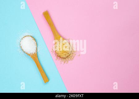 Weißer vs. Brauner Zucker, auf blauem und rosafarbenem Hintergrund. Teelöffel aus Holz voller Zucker, flach in Nahaufnahme. Stockfoto