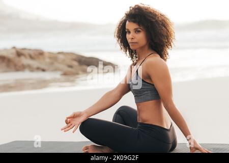 Junge Frau meditiert in Sportkleidung, während sie am Strand bei Sonnenuntergang sitzt Stockfoto