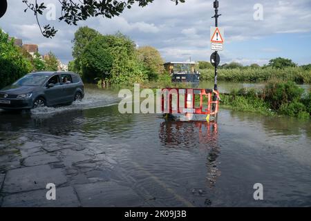 London, England, Großbritannien. 10.. September 2022. Auto überquert überflutete Straße am Ufer der Themse in Chiswick, West London. Der Mann schaut vom Autofenster auf das Wasser, das vom Bürgersteig sprudelt. Cristina Massei/Alamy Live News Stockfoto