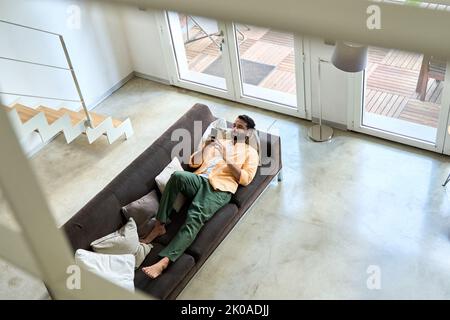 Entspannter indischer Mann, der zu Hause mit dem Handy auf der Couch chillt. Draufsicht Stockfoto