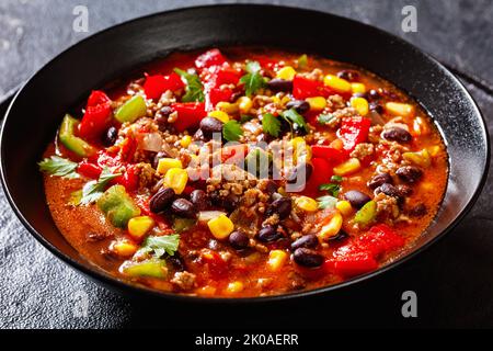 taco-Suppe aus Hackfleisch, Tomaten, gehackten grünen Chilis, Zwiebeln, Mais, Rote Bohnen und Taco-Würze in schwarzer Schüssel, Nahaufnahme Stockfoto