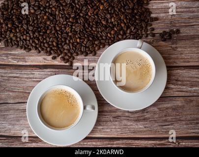Draufsicht auf zwei weiße Tassen mit Kaffeemilch auf Holztisch. Kaffeebohnen im Hintergrund Stockfoto