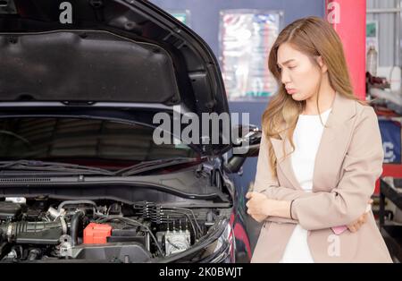 Junge asiatische Frau, die sich aufgeregt fühlt, während Notpanne Auto in der Garage, Emotion und Ausdruck, Kunde unglücklich und besorgt über Auto reparieren und wieder Stockfoto