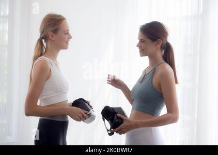 Zwei junge Frau, die eine VR-Brille mit Training und Bewegung trägt und sich im Wohnzimmer, in der virtuellen Realität und im Sport zum Üben des Workouts unterhält, li Stockfoto