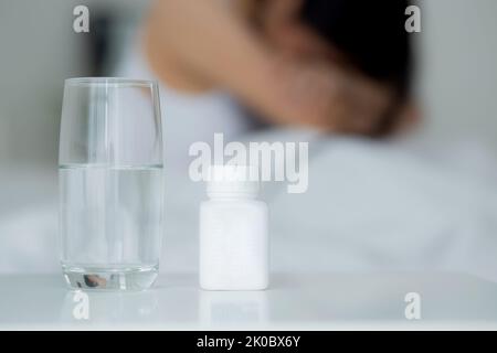 Junge asiatische Frau ist Depression und mit psychischen Problemen auf dem Bett sitzen, während Drogen und Wasser auf dem Schreibtisch im Schlafzimmer, Emotion und negativ, wom Stockfoto