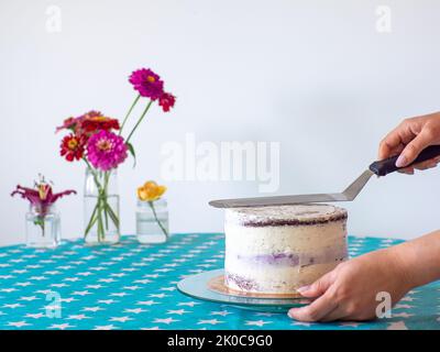 Die Creme mit einem Teigschaber von den Händen abgleichen und den runden Kuchen bedecken Stockfoto