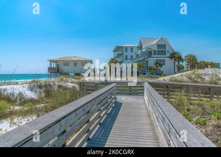 Blick auf Strandhäuser von einer Holzpromenade an einem Strand in Destin, Florida. Es gibt Gräser in der Nähe des Weges und einen Blick auf Strandhäuser in der Nähe der blu Stockfoto