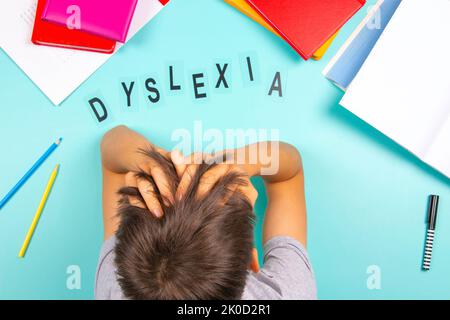 Traurig müde frustrierter Teenager, der mit vielen Büchern und Schulmaterial am Tisch saß. Wort Dyslexie auf hellblauem Hintergrund. Legasthenie, Lernen Stockfoto