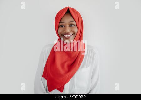 Porträt einer glücklichen afroamerikanischen muslimischen Frau in Hijab, die auf die Kamera schaut und lächelt, heller Studiohintergrund Stockfoto