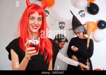 Die junge Frau, die als Teufel gekleidet war, lächelte mit einem roten Cocktail auf der Halloween-Party. Stockfoto