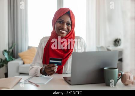 Bargeldlose Zahlung. Glückliche muslimische schwarze Frau in Hijab hält Kreditkarte und mit Laptop, Online-Shopping von zu Hause aus Stockfoto