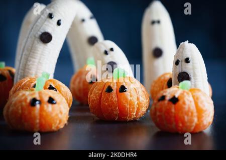 Halloween niedlichen Kürbis orange Frucht mit gruseligen Banane Geister Monster mit Schokolade Gesichter hinter ihnen. Gesunder Dessertjause mit lustigen Gesichtern. Stockfoto