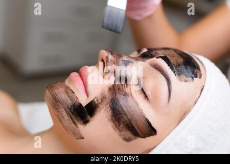 Kosmetologe, der schwarze Maske auf hübsches Frauengesicht mit schwarzen Handschuhen aufträgt, wunderschöne Frau im Spa, die Gesichtsbehandlungen hat Stockfoto