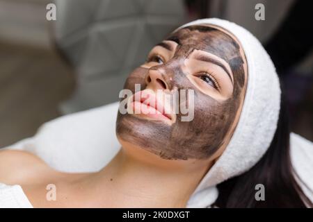 Kosmetologe, der schwarze Maske auf hübsches Frauengesicht mit schwarzen Handschuhen aufträgt, wunderschöne Frau im Spa, die Gesichtsbehandlungen hat Stockfoto