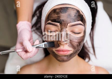 Kosmetologe, der schwarze Maske auf hübsches Frauengesicht mit schwarzen Handschuhen aufträgt, wunderschöne Frau im Spa, die Gesichtsbehandlungen hat Stockfoto