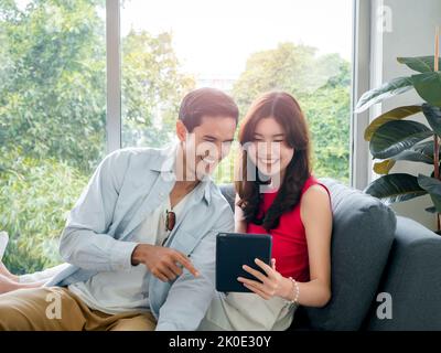 Glückliche junge schöne asiatische Frau und gutaussehende Mann lächeln zusammen, während sie auf Tablet-Bildschirm auf grauem Sofa in der Nähe des Glasfensters bei weißem Wohnzimmer ro Stockfoto