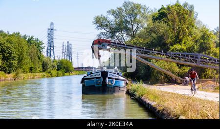 Eine motorisierte Barge, die auf einem Kanal festgemacht ist, wird in einem Sandsteinbruch mit Kies beladen, während ein Radfahrer an einem sonnigen Tag auf dem Schlepppfad vorbeifährt. Stockfoto