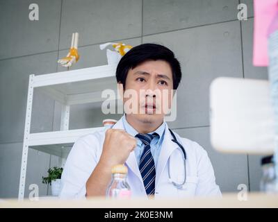 Porträt eines asiatischen männlichen Arztes in weißer Uniform mit einem Stethoskop, das auf dem Smartphone-Bildschirm auf die Kamera schaut, Online-Videoanruf und Ansporn für Pati Stockfoto