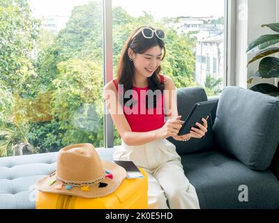 Junge schöne glückliche asiatische Frau mit Sonnenbrille mit digitalen Tablet-Computer mit Pass, Flugticket und Strandhut auf gelben Koffer, sitzen auf Stockfoto