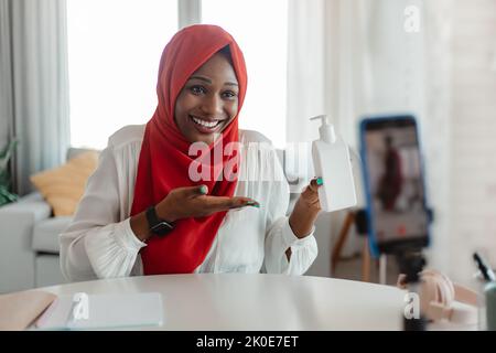 Glückliche schwarze muslimische Bloggerin, die Kosmetik zeigt und Videos für soziale Netzwerke auf dem Handy dreht Stockfoto
