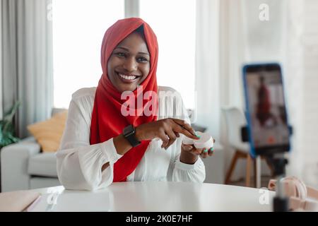 Glückliche afroamerikanische islamische Bloggerin, die Hautpflegeprodukte der Handykamera zeigt und Videos für ihren Beauty-Blog macht Stockfoto
