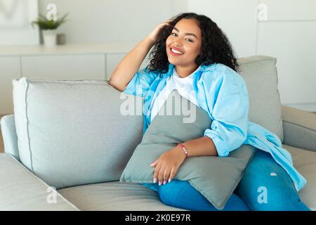 Happy African Lady Sitzt Auf Dem Sofa Und Hält Kissen Zu Hause Stockfoto