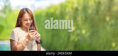 Asiatische Frau hält ein Glas Wasser aus dem Trinken auf Outdoor-Feld mit Bokeh grünen Banner Hintergrund. Stockfoto