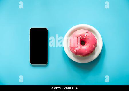 Donut mit pinker Vereisung und einem Smartphone auf blauem Hintergrund. Stockfoto