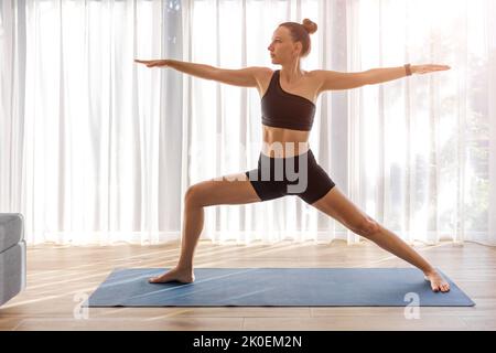 Junge Dame in schwarzer Sportkleidung, die im Zimmer mit großem Fenster Yoga macht Stockfoto