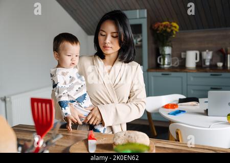 Glückliche Mutter hält ihren Sohn fest, während sie zu Hause Brot schneidet Stockfoto