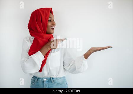 Überprüfen Sie dies. Glückliche afroamerikanische muslimische Frau im Hijab, die auf etwas in ihrer leeren Handfläche zeigt, Mockup Stockfoto
