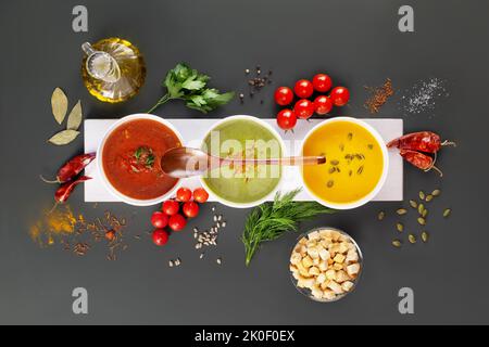 Set aus verschiedenen Gemüsecremesuppen mit Holzlöffel auf einem weißen Holzbrett mit Zutaten auf einem dunkelgrauen Hintergrund. Gemütliches Essen, vegetarische Küche Stockfoto